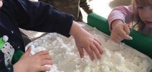 Children's hands playing with fluffy white material in a green bin.