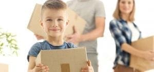 Child smiling while holding a cardboard box with parents in the background.