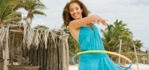 A woman in a blue dress hula-hoops on a beach with palm trees and a thatched hut.