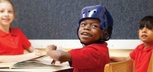 Young child in a blue protective helmet sits at a desk with two other children.