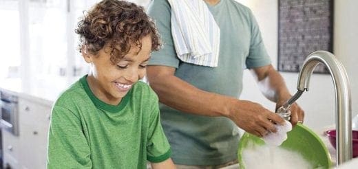 A child watches as an adult washes dishes in a kitchen sink.