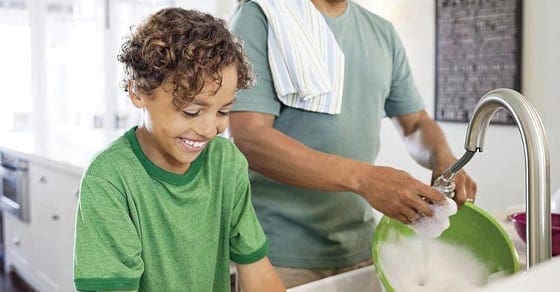 Child smiling while washing dishes with an adult in the background.