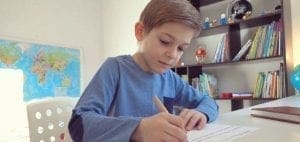 Boy in a blue shirt writing at a desk with books in the background