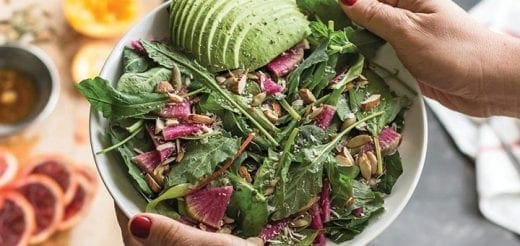 Salad with sliced avocado, radishes, and seeds being held by hands.
