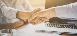 Two people shaking hands over a table with a laptop.