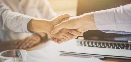 Two people shaking hands over a table with a laptop.