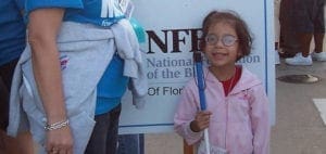 A young girl with glasses and a pink jacket smiles while holding a white cane.