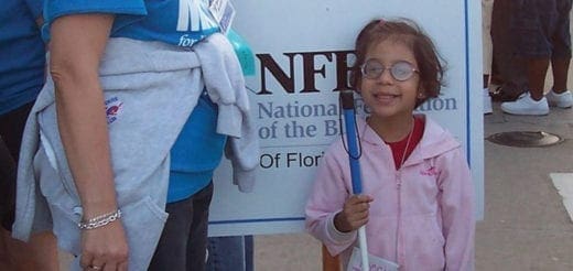 A young girl with glasses and a pink jacket smiles while holding a white cane.
