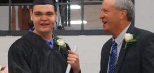 A graduate in a cap and gown holds a diploma while standing next to a man in a suit.