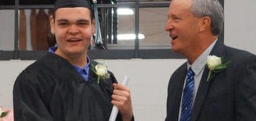 A graduate in a cap and gown holds a diploma while standing next to a man in a suit.