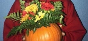 A carved pumpkin holds a bouquet of yellow and red flowers and green foliage.
