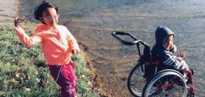 Two children are outdoors, one standing and one in a wheelchair near water.