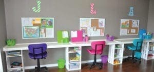 A long white desk with three workstations, each with a colorful chair, organized with bins and corkboards on the wall.