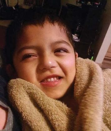 A young boy with dark hair smiles, wrapped in a tan towel.