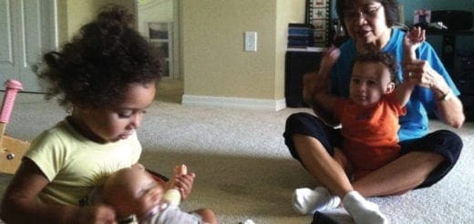 A young girl plays with a doll while an adult holds a baby, both sitting on the floor.
