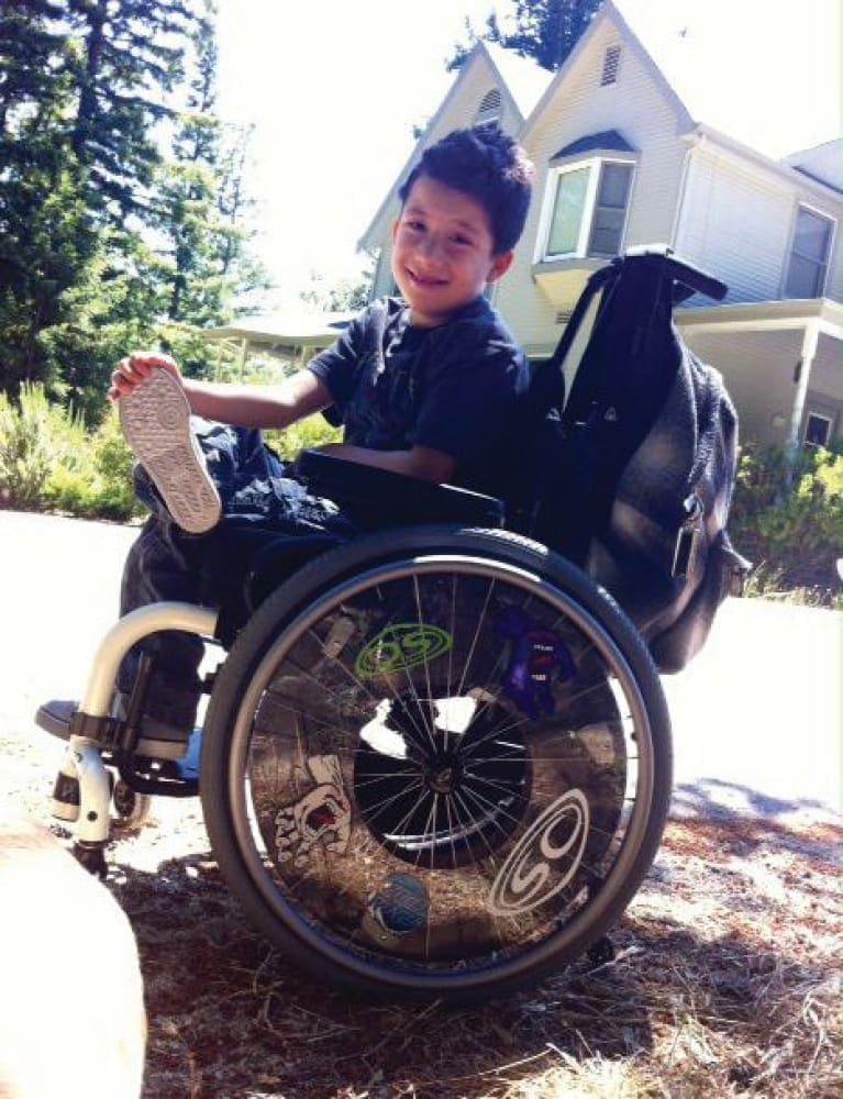 Joshy in his wheelchair with a house and trees in the background.