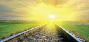 Train tracks lead toward a bright, sunlit horizon under a cloudy sky.
