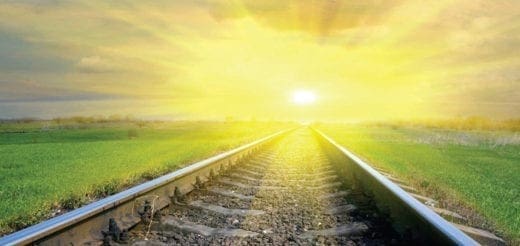 Train tracks lead toward a bright, sunlit horizon under a cloudy sky.