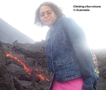 A woman wearing a denim jacket and pink skirt stands near a lava flow on a volcano.