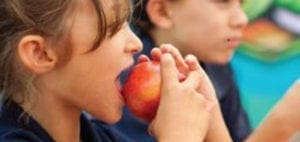 Child biting into a red apple while another child looks on.