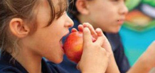 Child biting into a red apple while another child looks on.