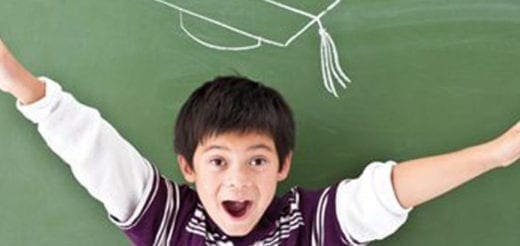 A young boy with arms outstretched in front of a chalkboard with a graduation cap drawn on it.