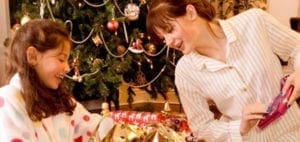 A young girl and a woman in pajamas look at presents under a Christmas tree.