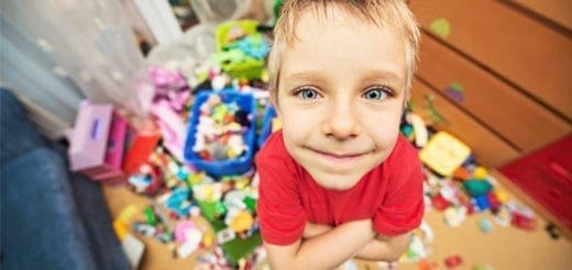 A young boy in a red shirt smiles with crossed arms in a messy room filled with toys.