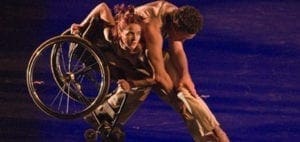 Dancers perform on a blue stage, with one dancer in a wheelchair being supported by another.