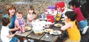 A group of children and an adult eat at a picnic table with KFC buckets and cups.