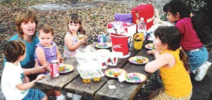 A group of children and an adult eat at a picnic table with KFC buckets and cups.