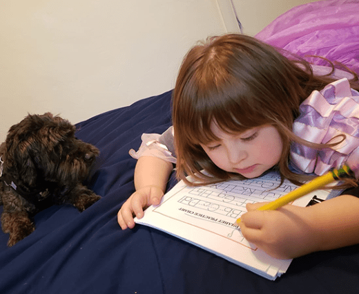 Child lying on a bed practicing handwriting on a worksheet with a small dog watching.