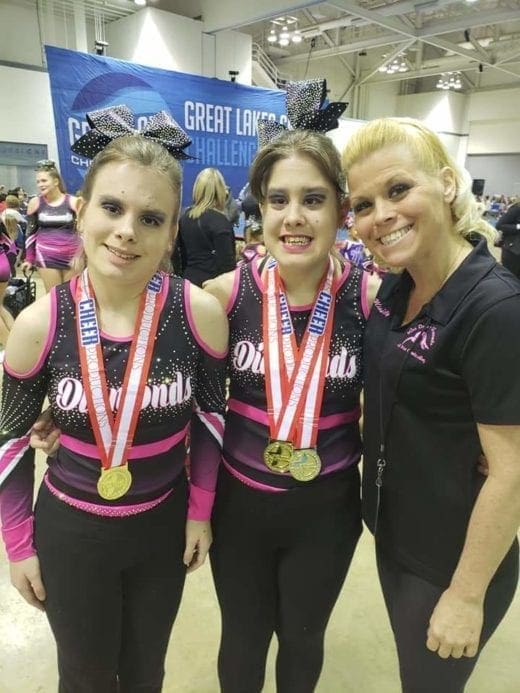 Kelly Long standing with her twin daughters, Kamryn and Reygan, wearing matching cheerleading uniforms and medals.