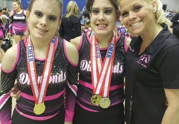 Kelly Long standing with her twin daughters, Kamryn and Reygan, wearing matching cheerleading uniforms and medals.