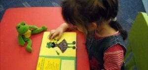 A child touches a picture of a frog in a book next to a stuffed frog.