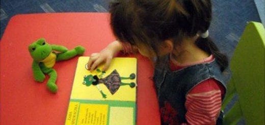 A child touches a picture of a frog in a book next to a stuffed frog.
