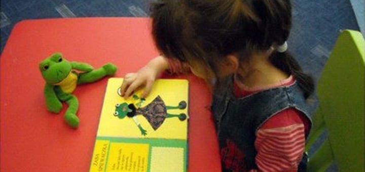 A child touches a picture of a frog in a book next to a stuffed frog.