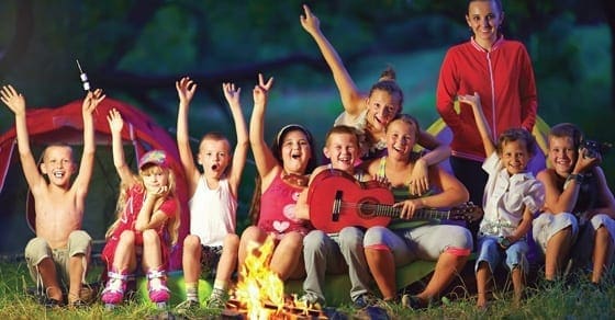 Children and adults smile and wave with their hands in the air around a campfire with tents in the background.