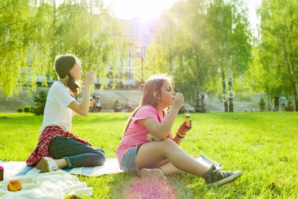 Two girls blow bubbles while sitting on grass in a park on a sunny day.