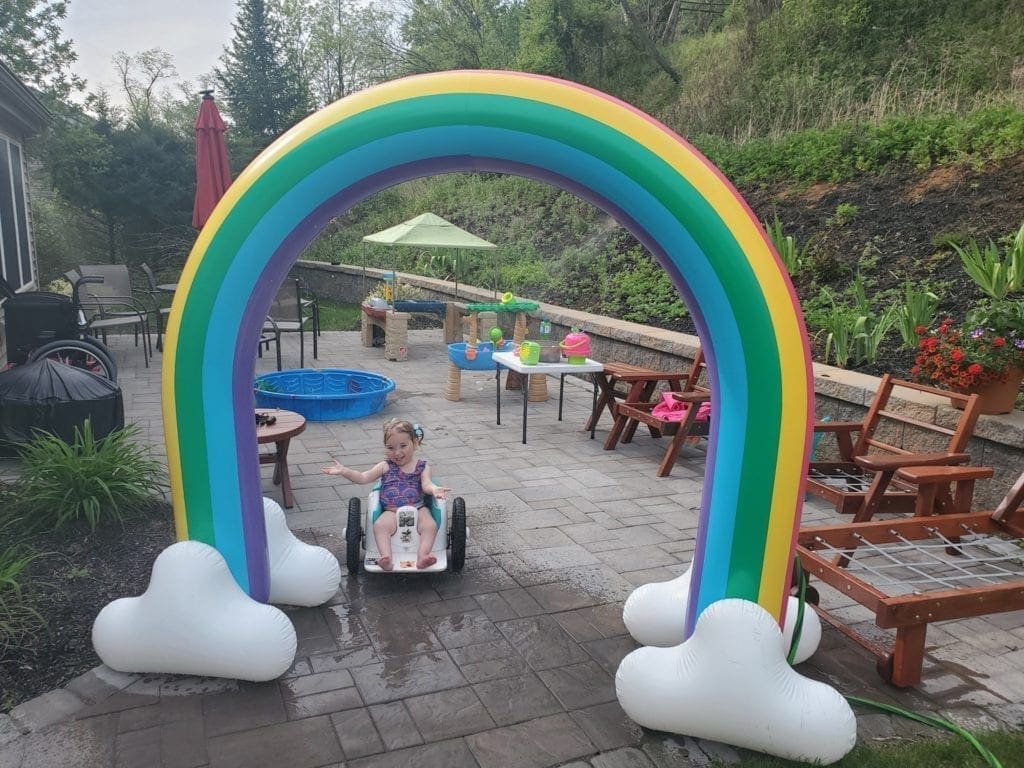 Child in a wheelchair playing under a rainbow sprinkler arch in a backyard.