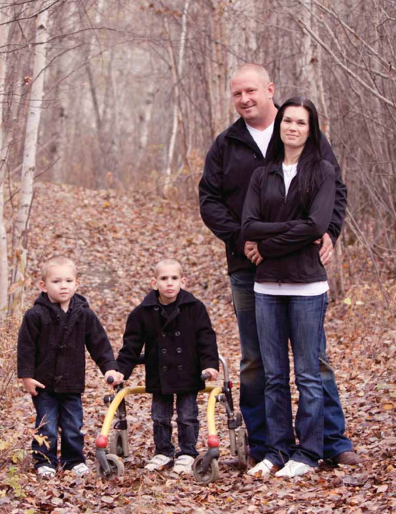 A family of four stands on a leaf-covered path in a wooded area.