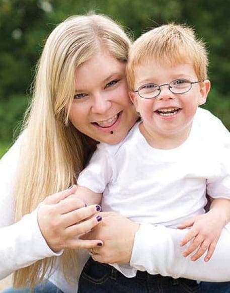 A smiling woman hugs a young boy wearing glasses.