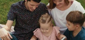 A family sits on a blanket outdoors, with parents looking down at their two young children.