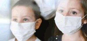 Two children wear white face masks and look out a window.