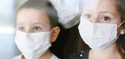 Two children wear white face masks and look out a window.