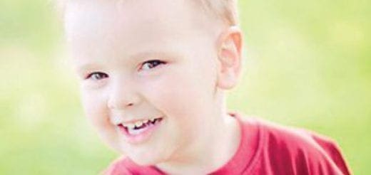 A young boy with blond hair wearing a red shirt smiles with his mouth open.