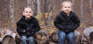Two young children sit on logs in front of a blurred background of trees.