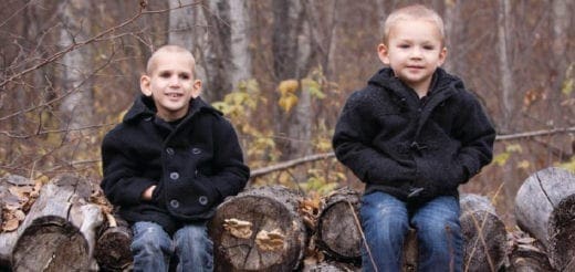 Two young children sit on logs in front of a blurred background of trees.