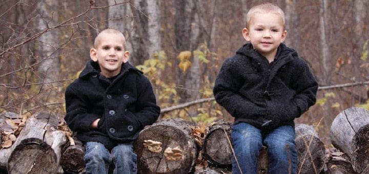 Two young children sit on logs in front of a blurred background of trees.