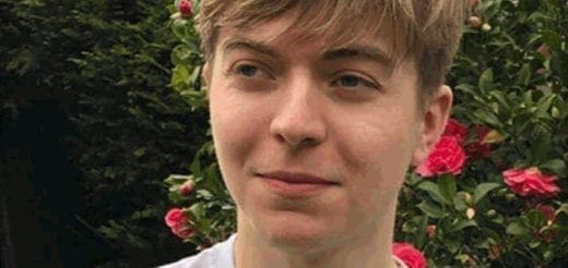 Young man with light brown hair and a white shirt, looking to his left with a bush of pink flowers behind him.